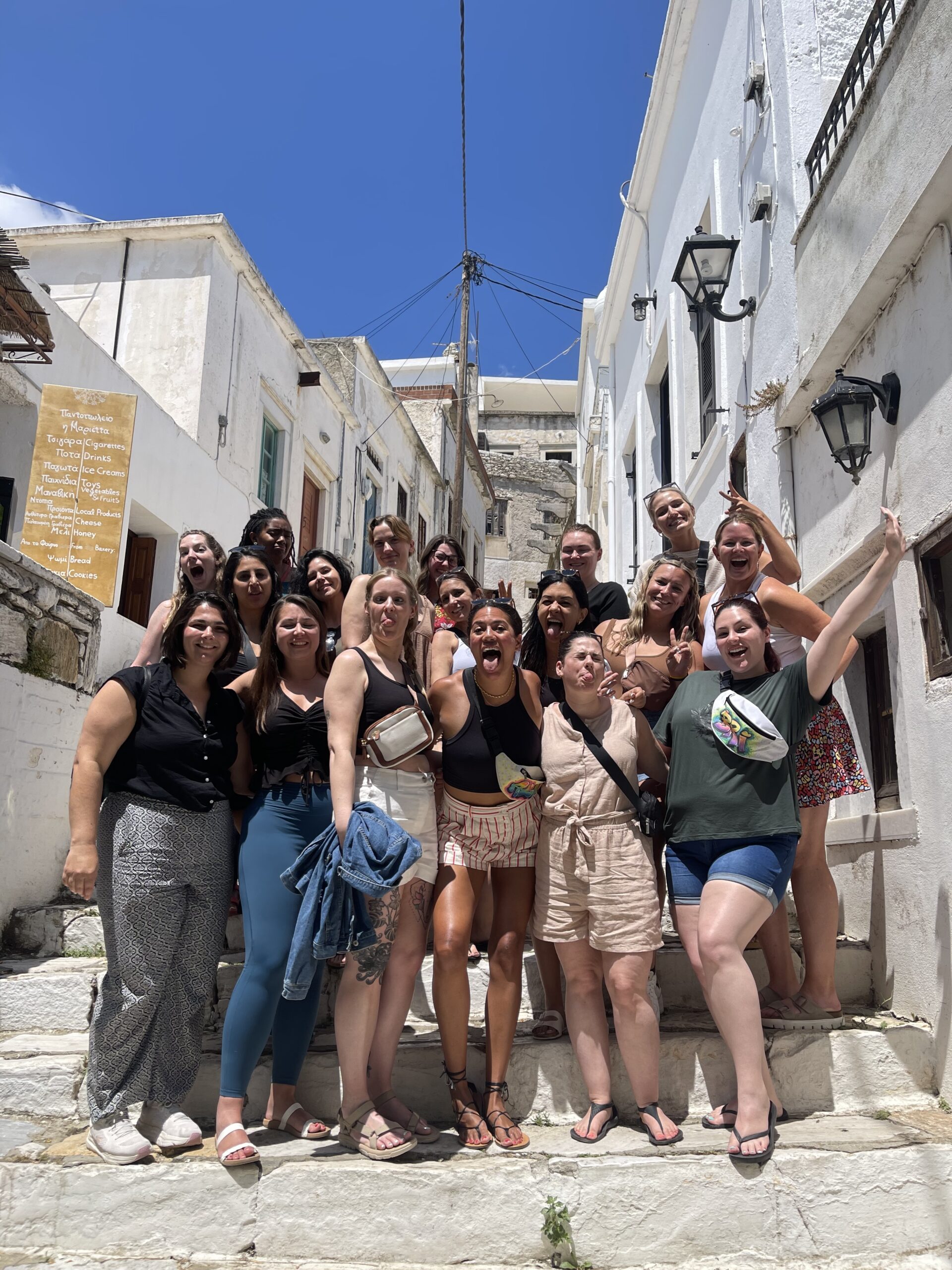 A large group of women posing and laughing together on white stone steps in a narrow alleyway of a Mediterranean village.