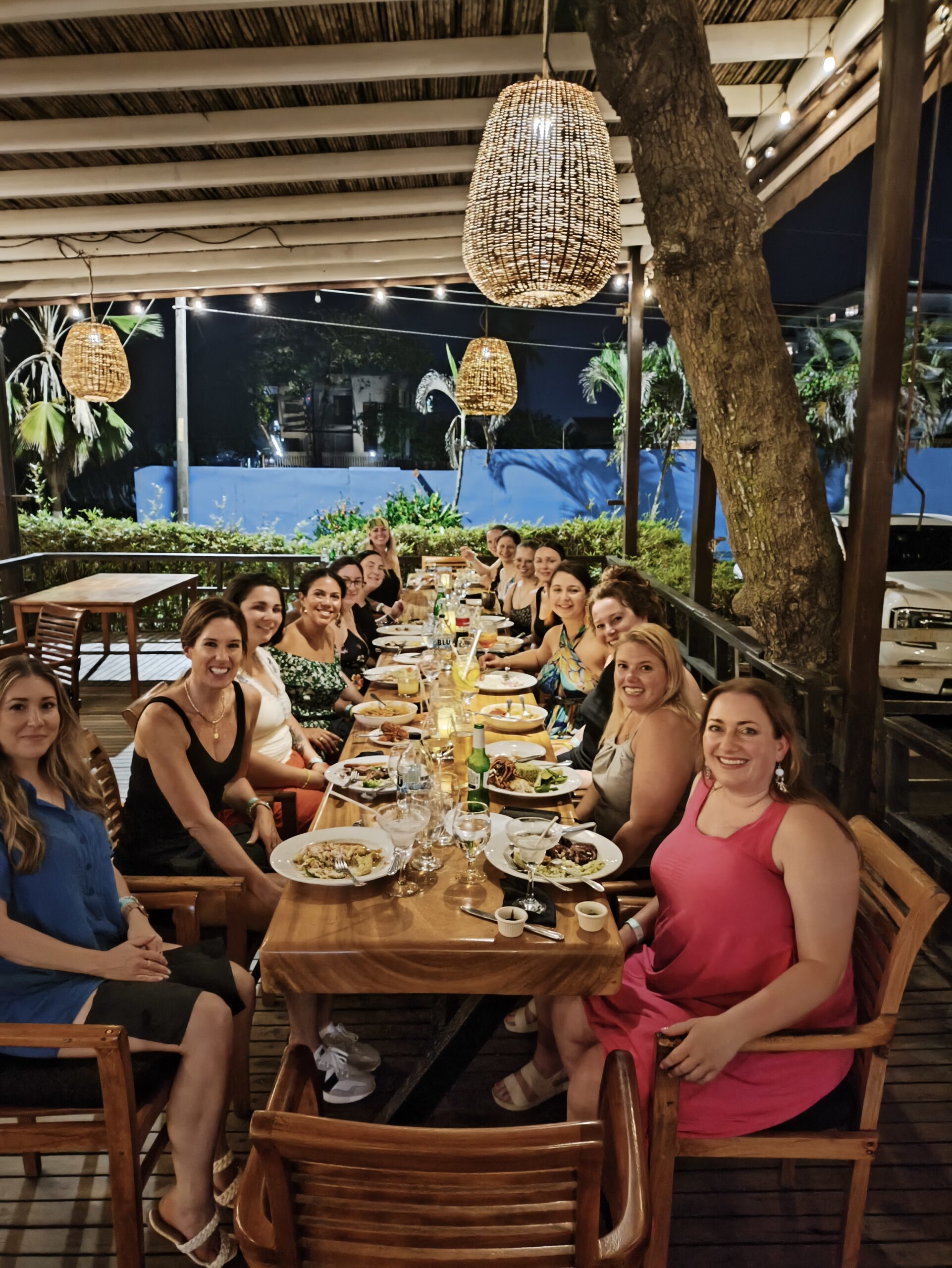 A group of women seated together at a long wooden dining table enjoying an outdoor dinner at night, surrounded by warm string lights and woven pendant lamps.