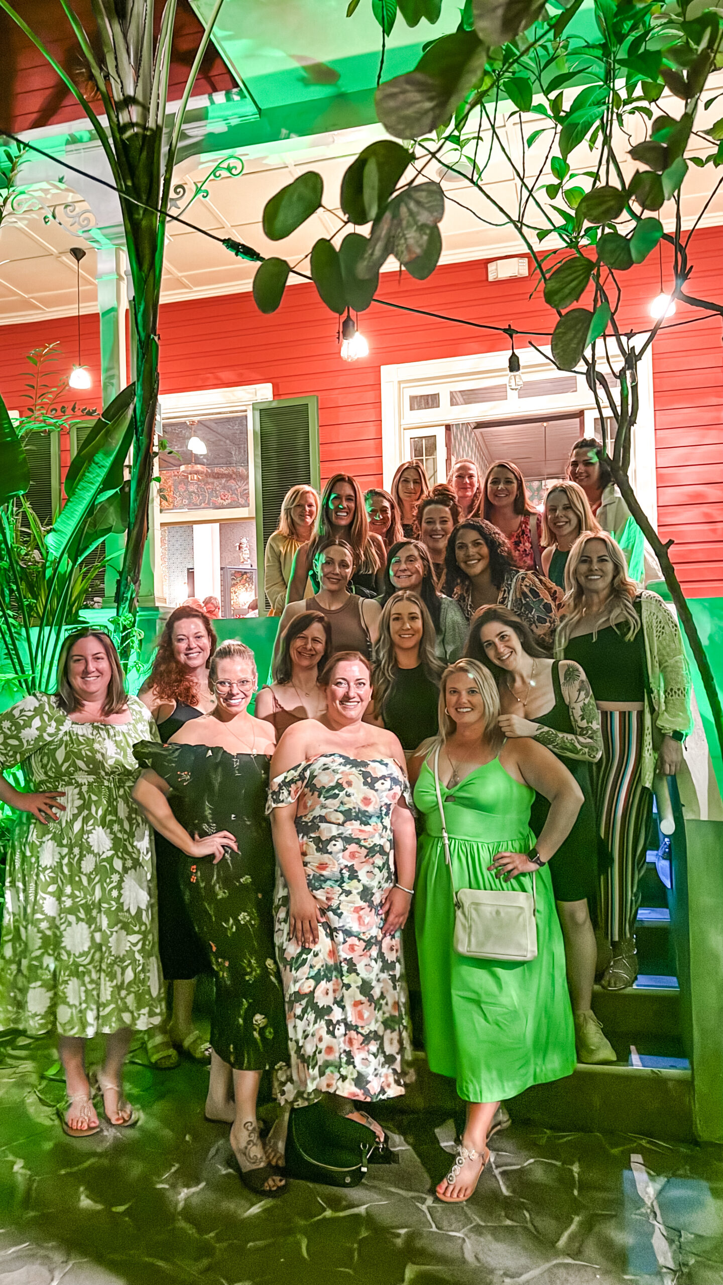 A large group of women dressed in shades of green posing together in front of a bright red building decorated with string lights.