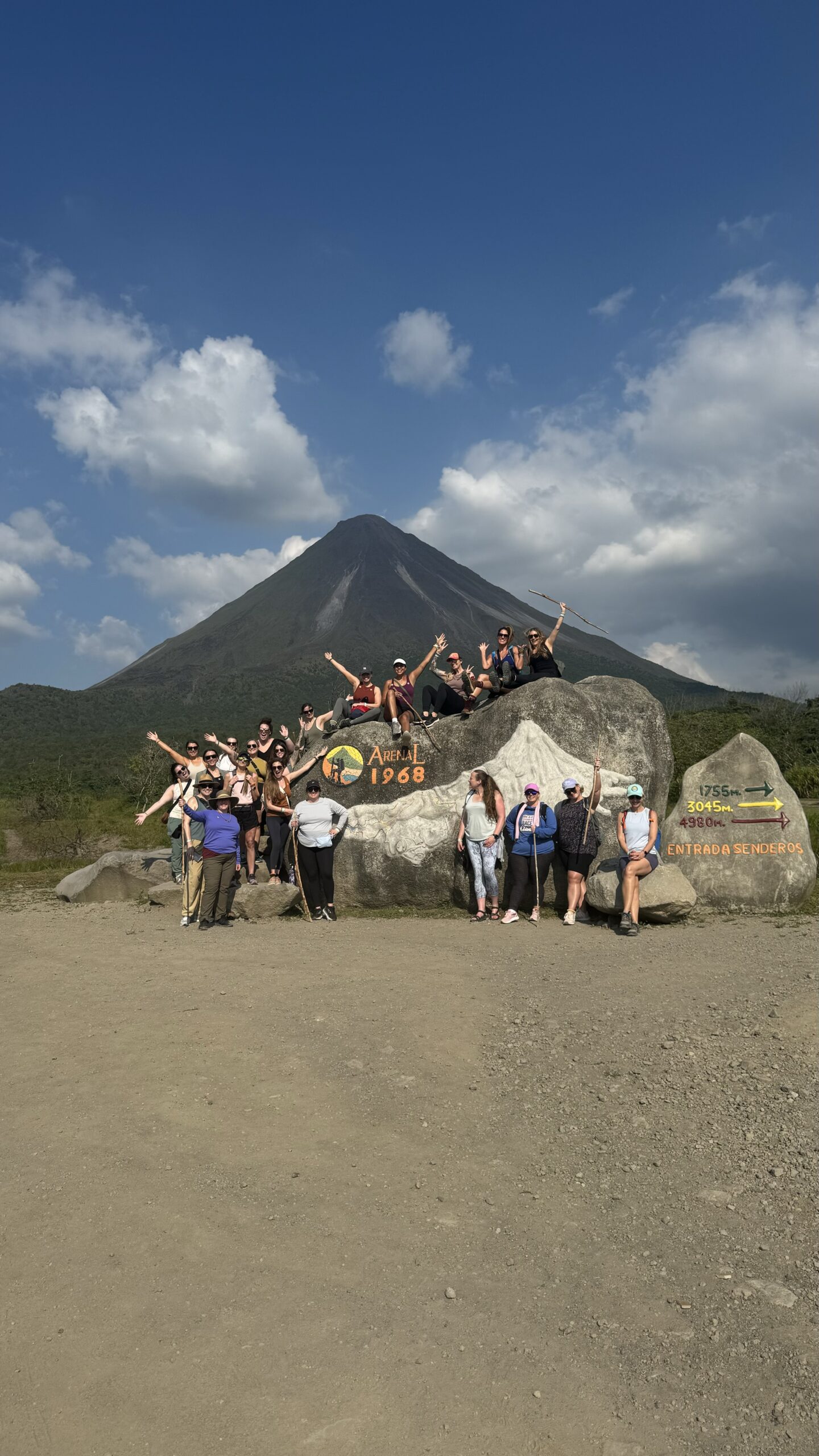 A large group of women posing with arms raised in front of a large volcano under a blue sky.