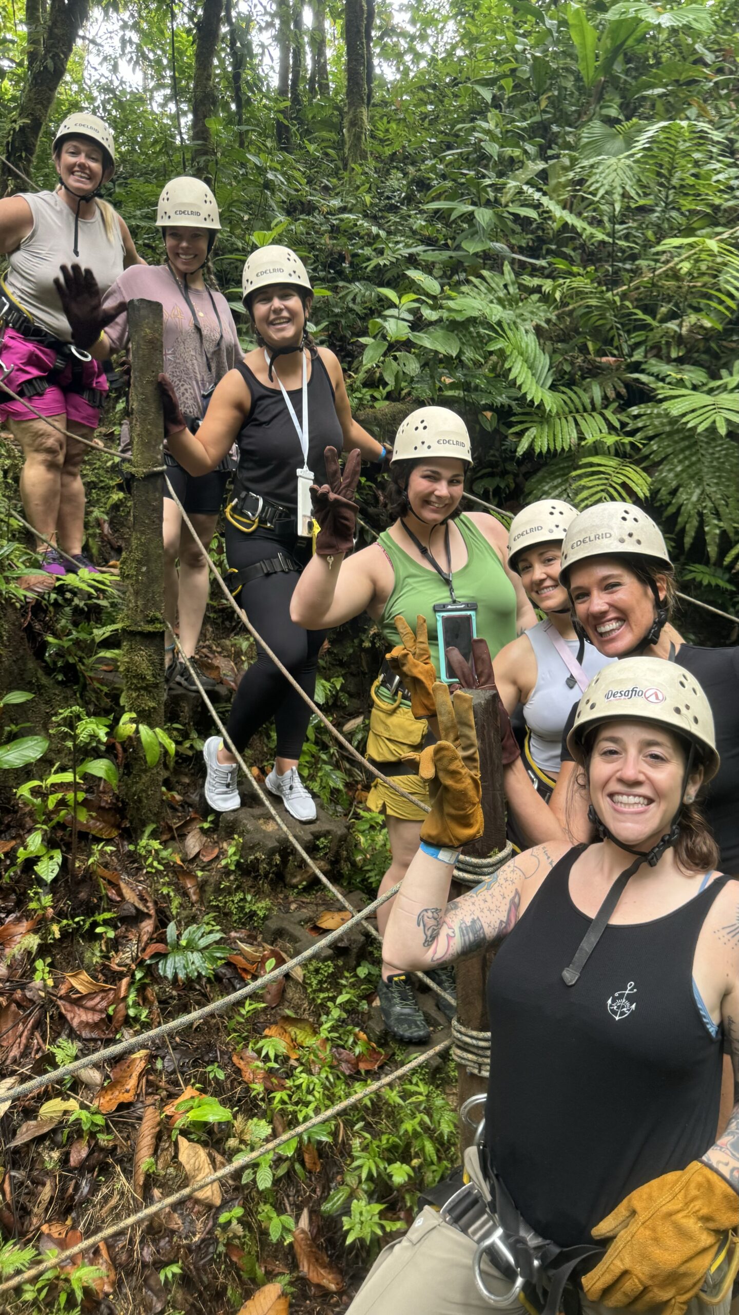 A woman taking a selfie while zip-lining through a lush jungle canopy, wearing a helmet, with friends visible behind her.