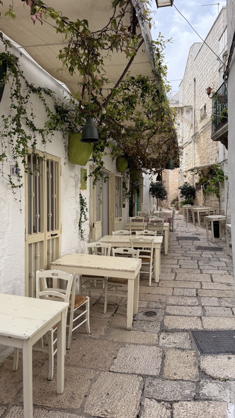 Narrow whitewashed alley in Cisternino, Puglia, with cream cafe tables and chairs set out under a vine-covered awning