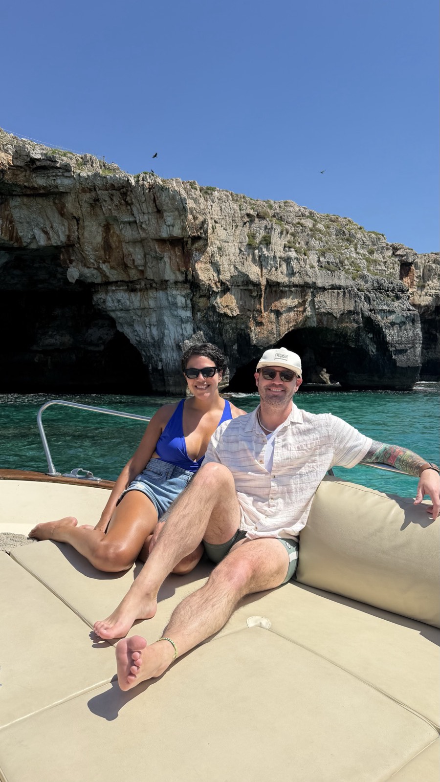 Ali and her partner sitting on the bow of a boat in turquoise water below limestone sea caves near Santa Maria di Leuca