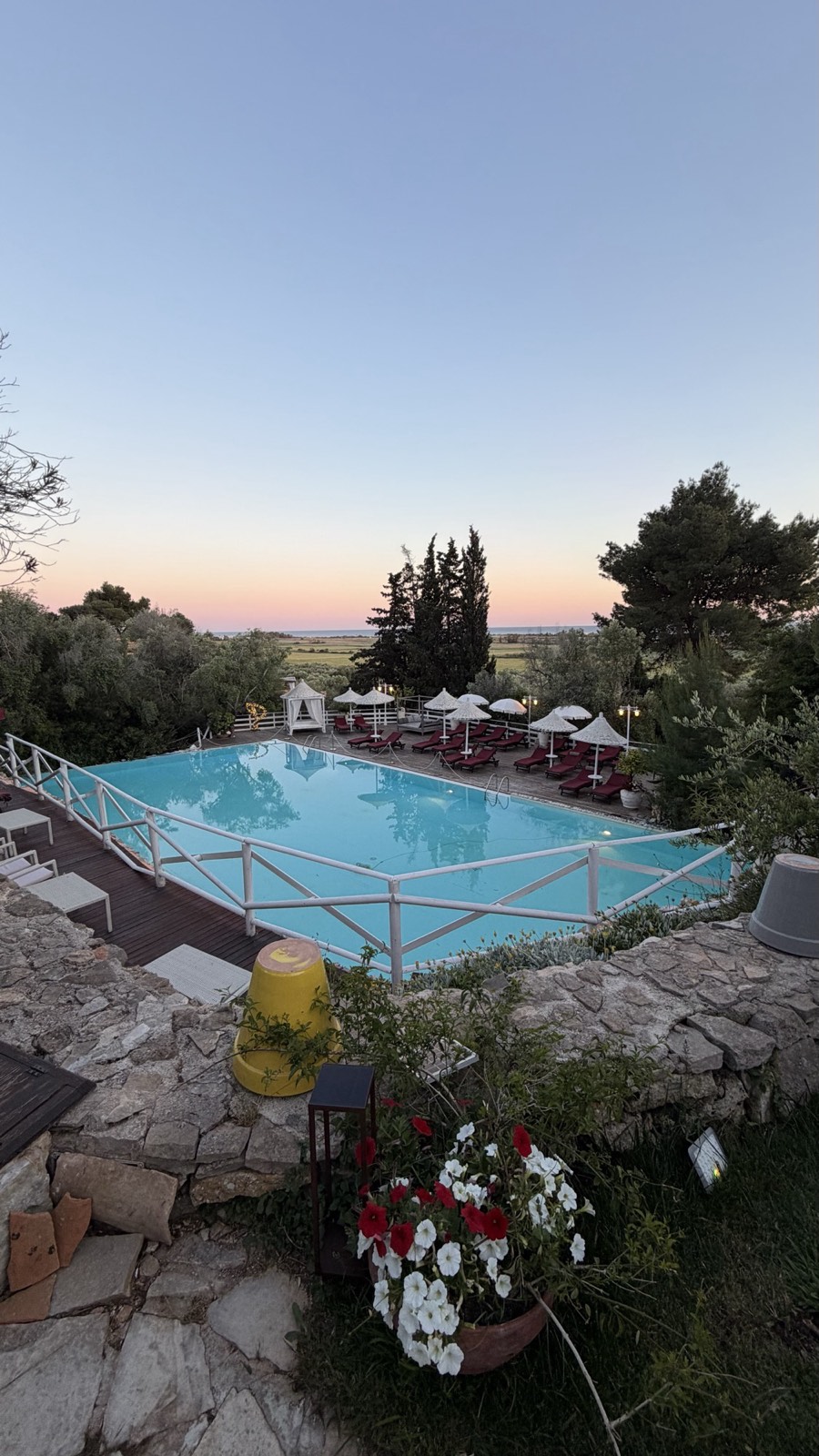 Rectangular pool at a Puglia masseria at dusk, lined with red loungers and straw parasols under a pink sky