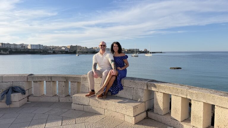 Ali and her partner sitting on a pale stone balustrade above the water in Otranto, Puglia, with the old town and moored sailboats behind them