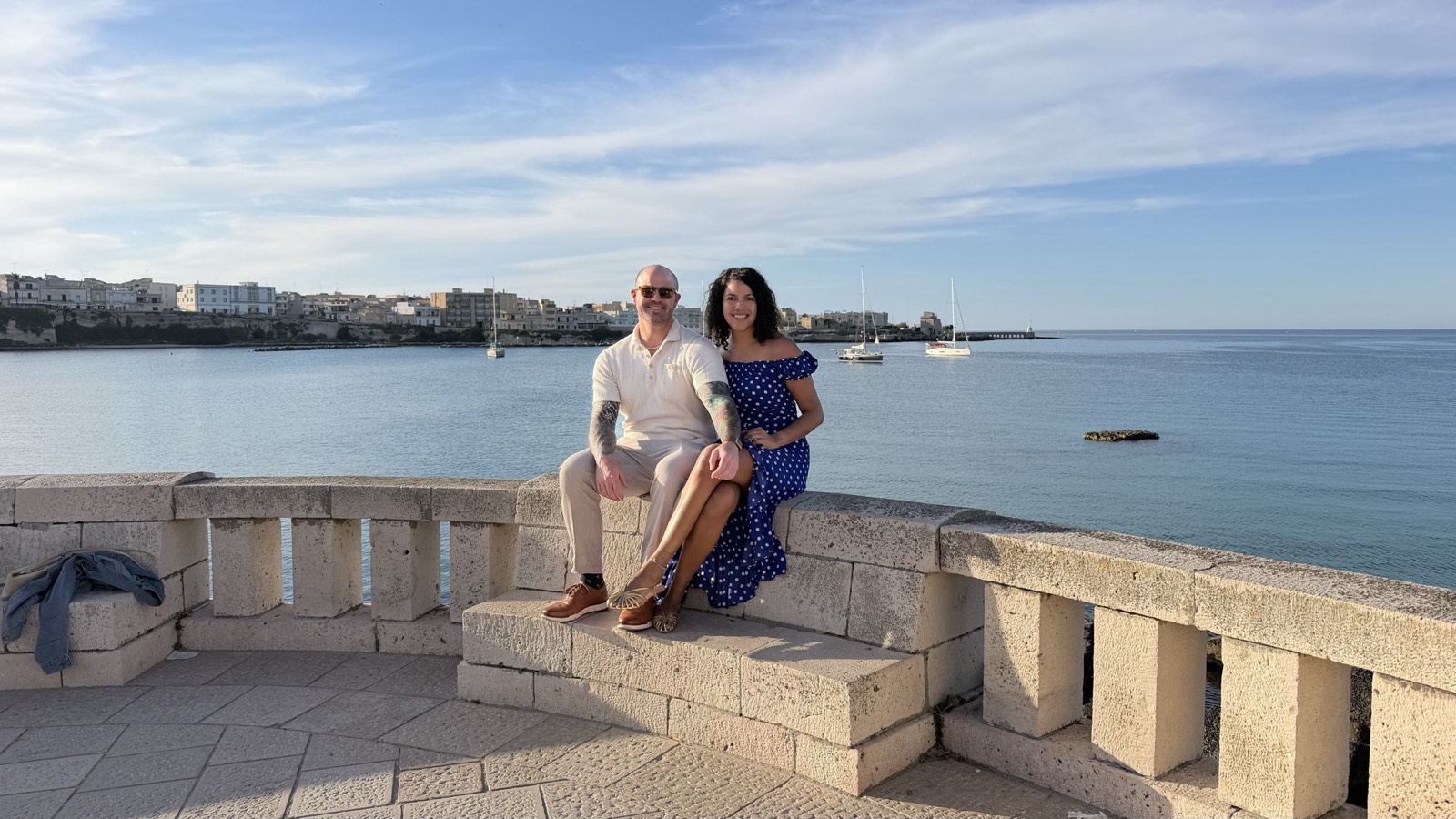Ali and her partner sitting on a pale stone balustrade above the water in Otranto, Puglia, with the old town and moored sailboats behind them